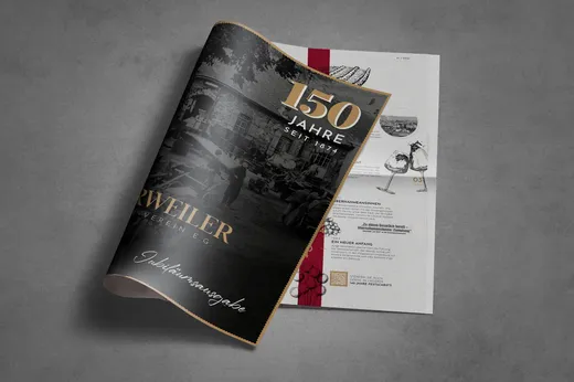 Auf einer grauen Fläche liegt eine aufgeschlagene Zeitschrift, deren Titelseite mit der Aufschrift „150 Jahre seit 1872“ in goldener Schrift und einer historischen Schwarz-Weiß-Fotografie verziert ist. Die Innenseite ist mit Grafiken und roten Akzenten gestaltet.