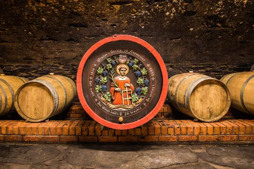 In einem Keller reihen sich hölzerne Weinfässer auf einer gemauerten Plattform aneinander. Das mittlere Fass ist mit einem kunstvollen Gemälde verziert, das einen Heiligen mit einem Kreuz in der Hand zeigt, umgeben von Weinreben und mit einer deutschen Inschrift versehen.
