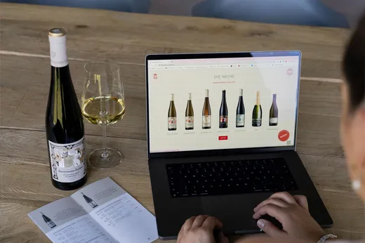 Eine Person surft auf einem Laptop auf der Website eines Weinhandels und sieht sich Weinflaschen an. Neben dem Laptop stehen auf einem Holztisch eine Weinflasche, ein gefülltes Weinglas und ein aufgeschlagener Katalog.