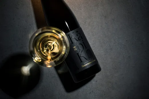 Ein Glas Weißwein, von oben betrachtet, neben einer dunklen Weinflasche mit der Aufschrift „Bassermann-Jordan“ auf einer grauen Fläche, wobei sowohl das Glas als auch die Flasche Schatten werfen.