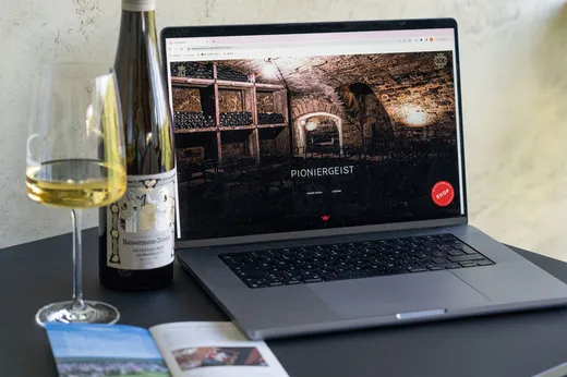 Auf einem Laptop, der auf einem Tisch steht, ist eine Website mit dem Bild eines Weinkellers zu sehen. Neben dem Laptop stehen eine Flasche und ein Glas Weißwein sowie eine aufgeschlagene Zeitschrift oder Broschüre.