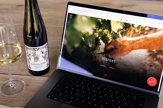 Ein Laptop, auf dem eine Website zu sehen ist, auf der eine Hand Trauben pflückt, daneben stehen ein Glas Weißwein und eine Flasche Riesling auf einem Holztisch.