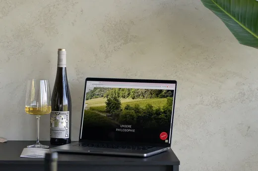 Auf einem schwarzen Tisch steht ein Laptop, auf dessen Bildschirm eine Weinbergszene zu sehen ist. Daneben stehen eine Flasche Weißwein und ein gefülltes Weinglas; in der Ecke liegt ein großes grünes Blatt, und im Hintergrund ist eine leicht strukturierte Wand zu sehen.