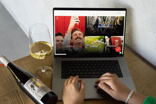 Eine Person nutzt einen Laptop, auf dem eine Website mit Bildern zum Thema Wein angezeigt wird. Neben dem Laptop stehen ein Glas Weißwein und eine Weinflasche auf einem Holztisch.