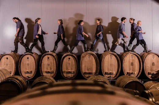 Sechs Personen gehen in einem Weingut auf großen Holzfässern in entgegengesetzte Richtungen und werfen dabei deutliche Schatten auf die Wand hinter sich. Im Vordergrund stehen weitere Fässer auf dem Boden.