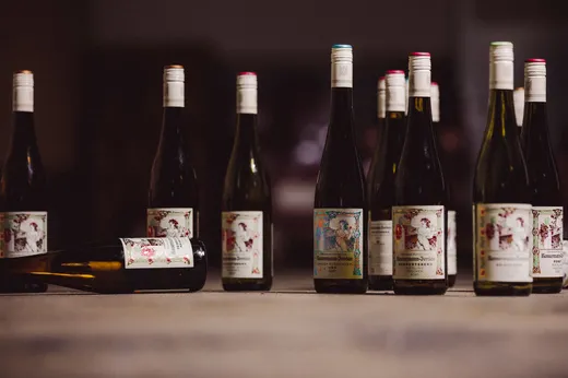 Mehrere Weinflaschen mit illustrierten Etiketten stehen aufrecht auf einer Holzfläche, während eine Flasche im Vordergrund horizontal liegt. Der Hintergrund ist unscharf und schwach beleuchtet.