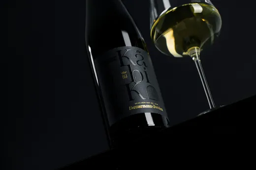 Eine Nahaufnahme einer dunklen Weinflasche mit der Aufschrift „Bassermann-jordan“ in goldener Schrift und einem teilweise sichtbaren Glas Weißwein vor schwarzem Hintergrund.
