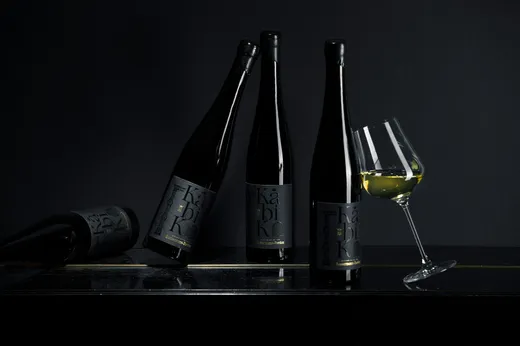 Vier dunkle Weinflaschen mit schwarzen Etiketten stehen auf einer schwarzen Fläche vor einem schwarzen Hintergrund; eine Flasche liegt auf der Seite, und neben den Flaschen steht ein geneigtes, mit Weißwein gefülltes Weinglas.
