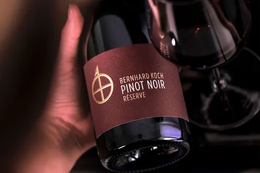Eine Hand hält eine Flasche „Bernhard Koch Pinot Noir Réserve“ mit einem dunkelbraunen Etikett neben einem teilweise gefüllten Weinglas.