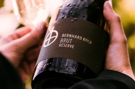 Nahaufnahme von Händen, die eine dunkle Flasche mit der Aufschrift „Bernhard Koch Brut Réserve“ halten; auf der Flsche sind Wassertropfen zu sehen, und der Hintergrund ist unscharf, was auf eine Außenaufnahme hindeutet.