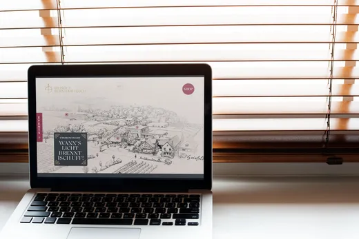 Auf einem Laptop, der auf einem weißen Tisch steht, ist eine Website mit einer Schwarz-Weiß-Illustration und Text zu sehen. Hinter dem Laptop befinden sich teilweise geschlossene Holzjalousien, durch die sanftes Licht hereinfällt.