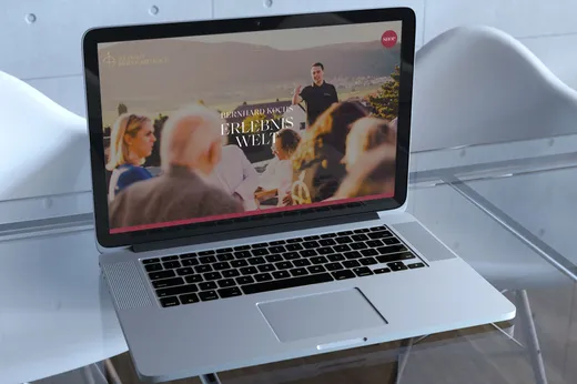 Auf einem Laptop, der auf einem Glastisch steht, ist eine Website zu sehen, auf der eine verschwommene Gruppe von Menschen und ein Mann zu sehen sind, der im Freien spricht; im Hintergrund stehen zwei weiße Stühle.