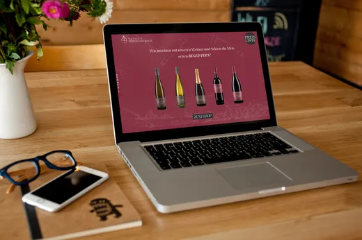 Auf einem Laptop, der auf einem Holztisch steht, ist eine Website mit fünf Weinflaschen zu sehen. Daneben liegen eine Brille und ein geschlossenes Notizbuch mit einem Stift. Die Szene spielt in einem gemütlichen, warm beleuchteten Raum.