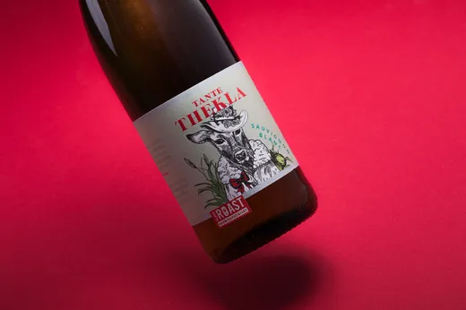 Eine Weinflasche mit einem „Tante Thekla“-Etikett, auf dem ein bekleidetes Reh vor einem leuchtend pink-roten Hintergrund abgebildet ist. Der untere Teil der Flasche ruht auf der Oberfläche.