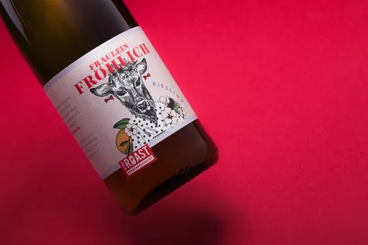 Eine Flasche mit dem Aufschrieb „Fräulein Fröhlich“, die schräg auf einem leuchtend roten Hintergrund steht. Das Etikett zeigt ein illustriertes Reh, Blumenmuster und eine auffällige rote Schrift.