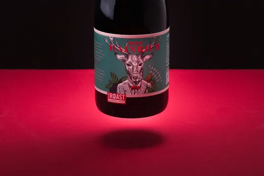 Eine Flasche Rotwein mit einem türkisfarbenen Etikett, auf dem ein Reh im Anzug abgebildet ist, vor einem schwarz-roten Farbverlaufshintergrund mit dramatischer Beleuchtung von unten.