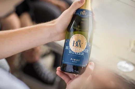 Eine Person hält eine Flasche Sekt mit der Aufschrift „Burkheimer Winzer Pinot Brut“ in der Hand. Das Etikett ist blau und goldfarben, und die Flasche wird in einer ungezwungenen, geselligen Atmosphäre präsentiert.