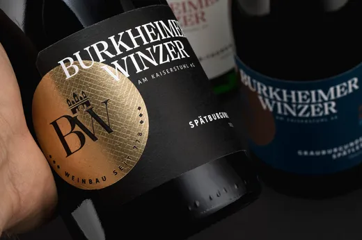Eine Nahaufnahme einer Hand, die eine dunkle Weinflasche mit der Aufschrift „Burkheimer Winzer“ hält, die mit einem gold-schwarzen Motiv verziert ist. Im Hintergrund sind weitere Weinflaschen mit ähnlichen Etiketten zu sehen.