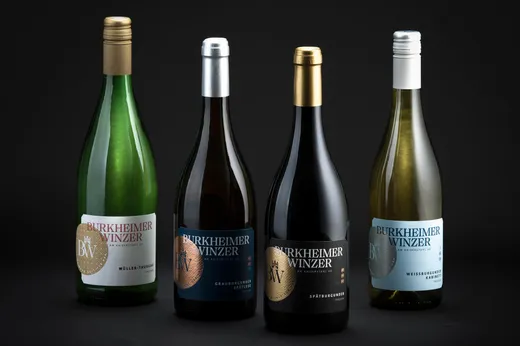 Vier Flaschen Burkheimer Winzer-Wein sind auf einem dunklen Hintergrund in einer Reihe angeordnet; jede Flasche hat ein eigenes Etikett und eine Folienkappe in Gold, Silber und Weiß, die die verschiedenen Weinsorten präsentieren.