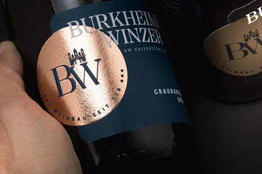 Eine Nahaufnahme einer Hand, die eine dunkle Weinflasche mit der Aufschrift „Burkheimer Winzer“ hält, versehen mit einem glänzenden Kupfersiegel, auf dem die Buchstaben „BW“ und ein kleines Schloss-Logo zu sehen sind. Im Hintergrund ist teilweise eine weitere Flasche zu erkennen.