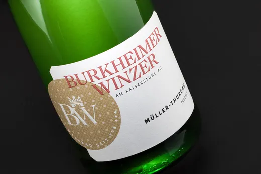 Eine Nahaufnahme einer grünen Weinflasche mit einem weißen Etikett, auf dem in brauner und schwarzer Schrift „Burkheimer Winzer Müller-Thurgau“ zu lesen ist, vor schwarzem Hintergrund.
