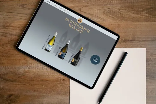 Auf einem Tablet, das auf einem Holztisch liegt, ist eine Webseite von „Burkheimer Winzer“ zu sehen, auf der drei Weinflaschen und eine Schaltfläche „Zum Shop“ abgebildet sind. Neben dem Tablet liegen ein schwarzer Stift und ein Blatt Papier.
