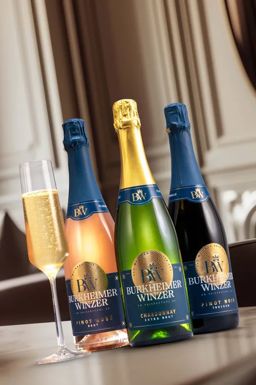 Drei Flaschen Burkheimer Winzer-Sekt (Pinot Rosé, Chardonnay und Pinot Noir) stehen auf einem Tisch neben einem gefüllten Sektglas in einem eleganten Innenraum.