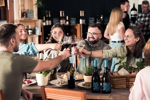 Fünf Personen sitzen um einen Tisch herum, lächeln und erheben ihre Weingläser zusammen. Auf dem Tisch stehen Flaschen, Pflanzen und Speisen. Im Hintergrund sind weitere Gäste sowie Regale mit Flaschen zu sehen.
