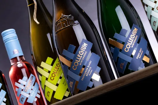 Nahaufnahme mehrerer Weinflaschen mit modernen, farbenfrohen Etiketten, auf denen „Collegium“ steht. Zu den Flaschen gehören Weißweine, Roséweine und Sekt, die schräg auf einer Holzkiste angeordnet sind.