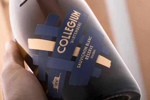 Eine Hand hält eine Weinflasche mit der Aufschrift „COLLEGIUM WIRTEMBERG SAUVIGNON BLANC RÉSERVE trocken“ und einem modernen blau-goldenen Design, das aus sich überlappenden Streifen und der Abbildung eines Gebäudes besteht.