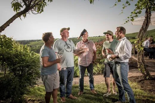 Fünf Männer stehen draußen in einem Garten, lachen und unterhalten sich. Ein Mann wirft eine Weinflasche in die Luft, während die anderen mit Weingläsern in der Hand zuschauen. Im Hintergrund sind grüne Hügel und Bäume unter einem klaren Himmel zu sehen.