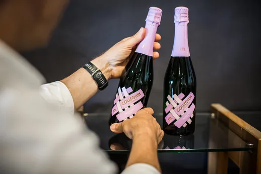 Eine Person in einem weißen Hemd hält eine Flasche Sekt mit der Aufschrift „Collegium“ und einem rosa Verschluss in der Hand, neben einer weiteren identischen Flasche auf einem Glasregal.