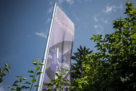 Eine hohe, senkrecht stehende Flagge mit der Aufschrift „COLLEGIUM“ und abstrakten Gebäudegrafiken ragt zwischen grünen, belaubten Ästen vor einem blauen Himmel mit vereinzelten Wolken empor.