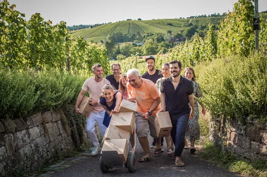 Eine Gruppe lächelnder Menschen, die Kisten tragen und einen Wagen schieben, geht an einem sonnigen Tag gemeinsam einen Weg hinauf, der von üppig grünen Weinbergen gesäumt ist. Sanfte Hügel und Weinreben bilden den Hintergrund.