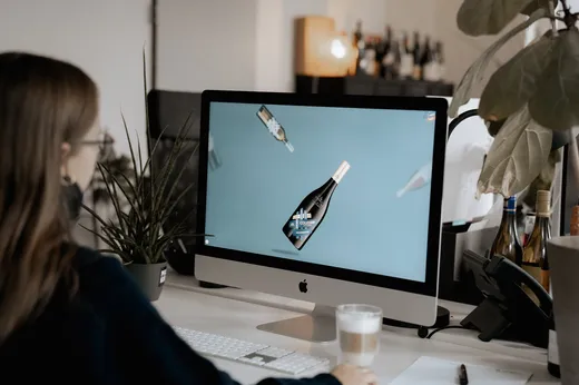 Eine Person sitzt an einem Schreibtisch mit einem Computer und betrachtet einen Bildschirm, auf dem schwebende Weinflaschen vor blauem Hintergrund zu sehen sind. Auf dem Schreibtisch stehen eine Tasse Kaffee, Pflanzen und Flaschen.
