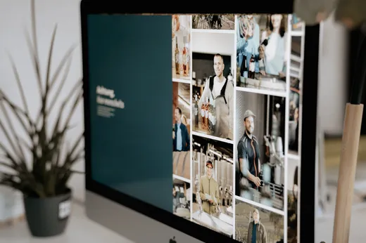 Auf einem Computerbildschirm ist eine Fotocollage mit verschiedenen Personen sowie ein dunkelblauer Bereich mit Text zu sehen; neben dem Monitor sind eine Topfpflanze und ein Holzgegenstand zu erkennen.