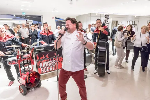 Ein Mann in einem weißen Hemd und einer roten Hose singt voller Leidenschaft vor einem begeisterten Publikum ins Mikrofon. Hinter ihm spielt eine Band mit Schlagzeug und Kontrabass, und das Publikum klatscht und lächelt in einem hellen Innenraum.