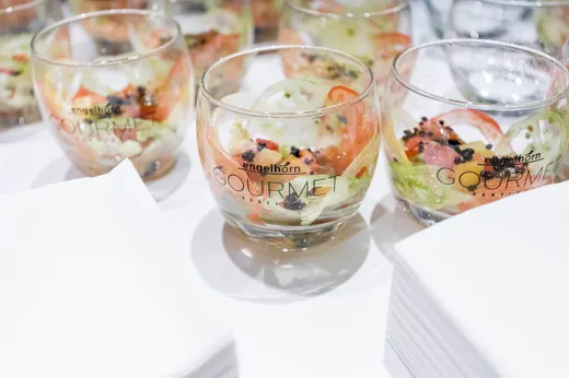 Mehrere durchsichtige Gläser mit der Aufschrift „engelhorn GOURMET“ sind mit einem Salat aus Tomaten, Gurken und Kräutern gefüllt und stehen auf einer weißen Unterlage neben einem Stapel weißer Servietten.