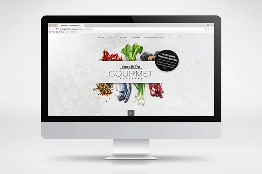 Auf einem Computermonitor ist die Website des Engelhorn-Gourmetfestivals zu sehen, auf der frisches Gemüse und Fisch vor dem Festival-Logo auf weißem Hintergrund angeordnet sind.