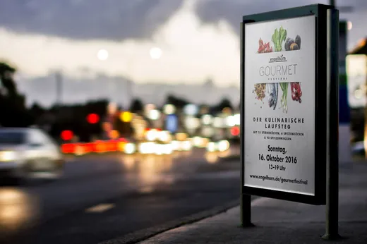 An einer Bushaltestelle in der Abenddämmerung ist ein Plakat für ein Gourmet-Festival zu sehen, auf dem Gemüse abgebildet ist und Veranstaltungsdetails stehen, während die Scheinwerfer der Autos auf der nahegelegenen Straße vorbeirauschen.