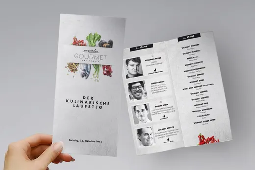 Eine Hand hält eine gefaltete Broschüre für ein Gourmet-Festival in der Hand. Auf dem Titelblatt sind buntes Gemüse und der Titel der Veranstaltung auf Deutsch abgebildet. Im Innenteil sind die Namen der Köche mit Schwarz-Weiß-Porträts sowie Details zur Veranstaltung aufgeführt.