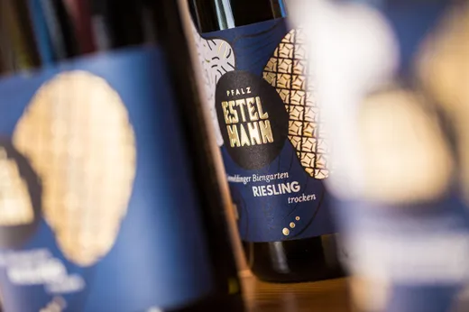 Nahaufnahme von blauen Weinetiketten mit goldenen kreisförmigen Mustern und der Aufschrift „Pfalz Estelmann Riesling trocken“, wobei einige Etiketten im Vordergrund und Hintergrund unscharf sind.