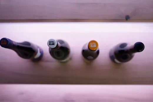 Vier Weinflaschen sind in einer Reihe angeordnet, von oben betrachtet. Jede Flasche hat einen farbigen Verschluss , auf denen jeweils „ESTELMANN“ steht. Der Hintergrund besteht aus sanft beleuchtetem Holz.