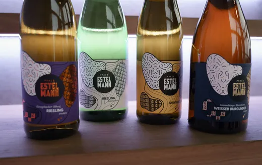 Auf einer Holzunterlage stehen vier Flaschen Estelmann-Wein, jede mit einem farbenfrohen, kunstvollen Etikett. Zu den Sorten gehören Riesling, Sauvignon und Weißer Burgunder. Im Hintergrund strahlt ein sanftes Licht.