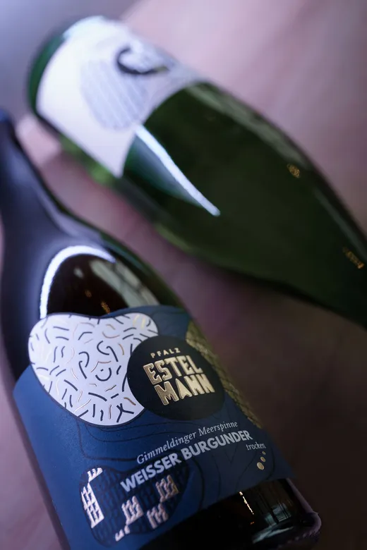 Zwei Weinflaschen liegen auf einer Holzfläche; die vordere Flasche hat ein blau-weißes, abstrakt gestaltetes Etikett mit der Aufschrift „Pfalz Estelmann Weißer Burgunder“, während die dahinter liegende Flasche ein weiß-grünes Etikett trägt.