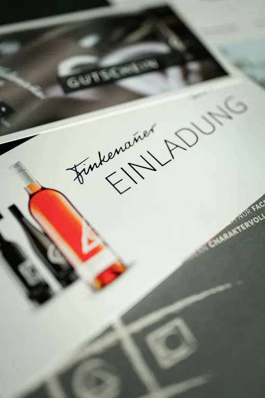 Eine Nahaufnahme einer Einladungskarte, auf der eine Flasche Roséwein und die deutschen Worte „Finkenauer EINLADUNG“ zu sehen sind. Im Hintergrund sind teilweise weitere, unscharfe Karten zu erkennen.