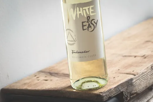 Eine Flasche Weißwein mit der Aufschrift „White &Easy; Finkenauer, Weißwein Cuvée“ steht auf einer rustikalen Holzunterlage, leicht nach rechts geneigt, wobei ein sanftes Licht das Etikett der Flasche beleuchtet.