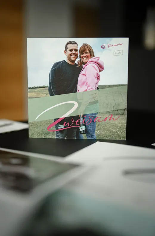 Eine Karte, auf der ein lächelnder Mann und eine lächelnde Frau gemeinsam im Freien stehen; auf der Vorderseite steht in rosa Schreibschrift das Wort „Zweisam“. Im Hintergrund sind eine Wiese und ein bewölkter Himmel zu sehen.