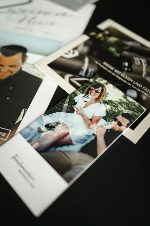 Eine Nahaufnahme von auf einem Tisch verstreuten gedruckten Fotos, auf denen eine lächelnde Frau mit Sonnenbrille und in einem weißen Kleid zu sehen ist, die mit einem Mann im Freien sitzt, sowie weitere Personen und teilweise sichtbare Einladungen zu Veranstaltungen.