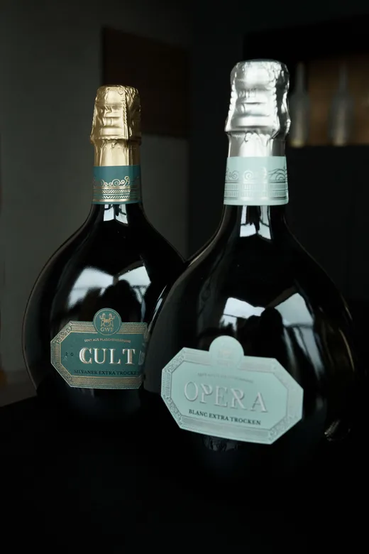 Zwei dunkle Glasflaschen mit Sekt, deren Verschlüsse mit Folie umwickelt sind – die eine mit der Aufschrift „CULT“ und mit Goldfolie, die andere mit der Aufschrift „OPERA“ und mit Silberfolie –, sind auf einer schwarzen Fläche vor einem unscharfen Hintergrund zu sehen.